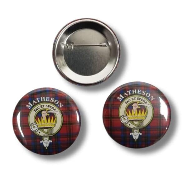 Three round badges from the Armstrong Clan collection feature a red and blue tartan pattern. Two of the badges display the Matheson crest, which includes a green shield with a flaming torch, symbolizing heritage associated with the Armstrong Clan. The third badge shows the back with a pin.