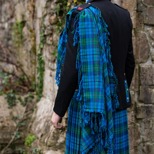 A man in a Welsh Tartan Medium Weight Premium Wool Fly Plaid kilt standing next to a stone wall.
