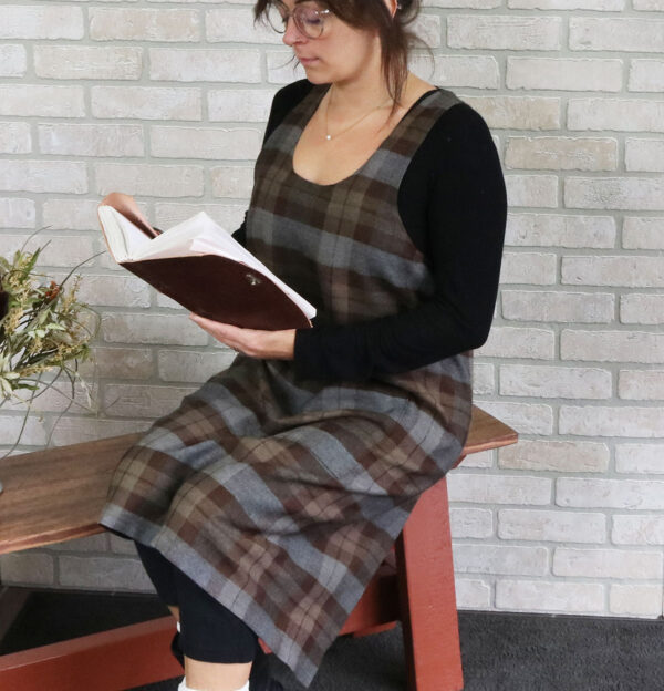 A woman wearing a Tartan Cross Back Apron - Homespun Wool Blend sits on a bench, engrossed in her book.