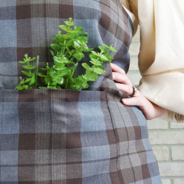 A woman wearing a Tartan Cross Back Apron - Homespun Wool Blend with a plant in the pocket.