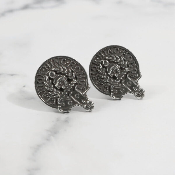 A pair of silver stud earrings on a marble surface, accompanied by Gaelic Themes Clan Crest Lapel Pins.