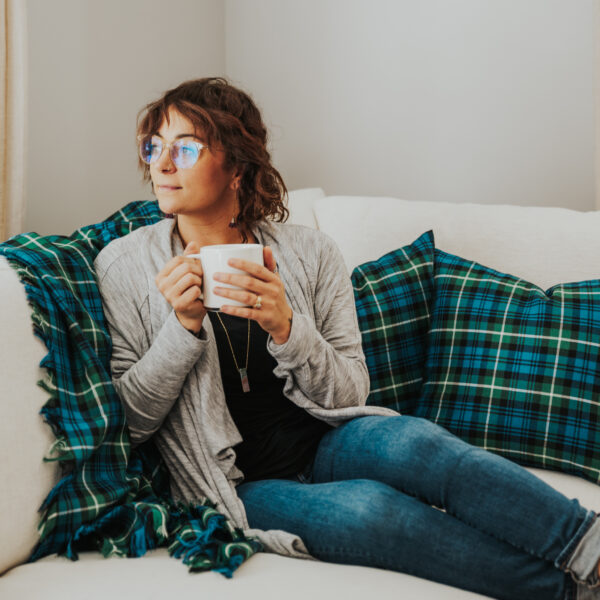 A woman sitting on a couch wrapped in a Homespun Tartan Blanket/Throw, holding a cup of coffee.
