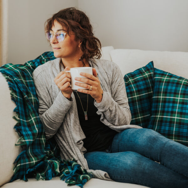 A woman sitting on a couch wrapped in a Homespun Tartan Blanket/Throw, holding a cup of coffee.