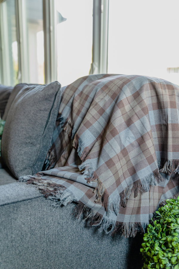 An OUTLANDER Throw Poly/Viscose Tartan draped stylishly on a couch, complemented by a plant in the background.