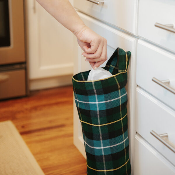 A person placing a green and black tartan trash bag in a kitchen cabinet using the Tartan Plastic Bag Holder - Homespun Wool Blend.