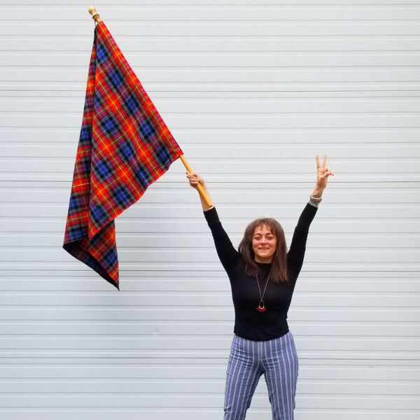 A woman proudly holding an LGBT Pride Tartan Flags - Homespun Wool Blend in front of a garage.