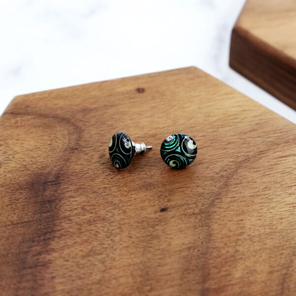 A pair of black and green Triskelion Spiral Paua Shell earrings on a wooden board.