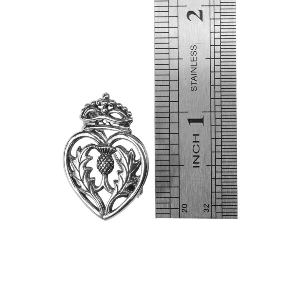 A silver heart shaped locket, adorned with a Luckenbooth Thistle Brooch, next to a ruler.