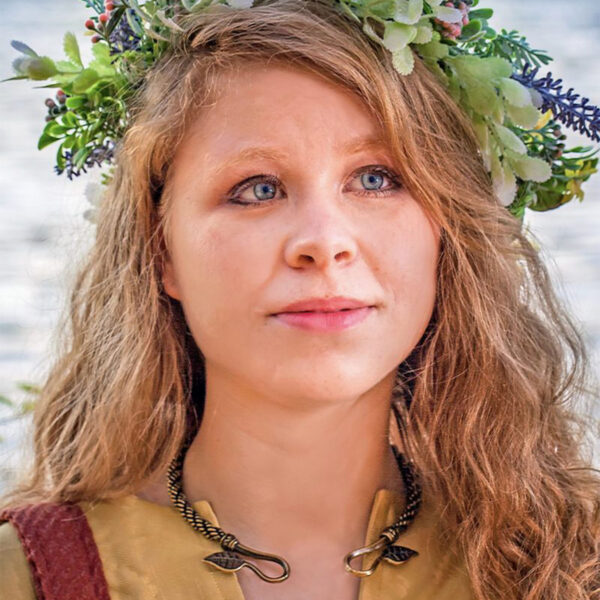A girl wearing a flower crown in front of a body of water, adorned with an Ash Leaf Torc Medium Braid.