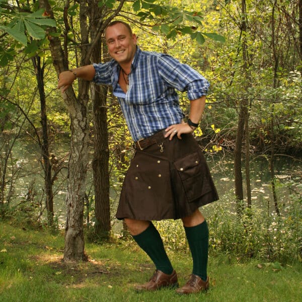 In the Wilderness Kilt - Free Kilt Hanger, a man effortlessly stands against a tree in his traditional kilt.