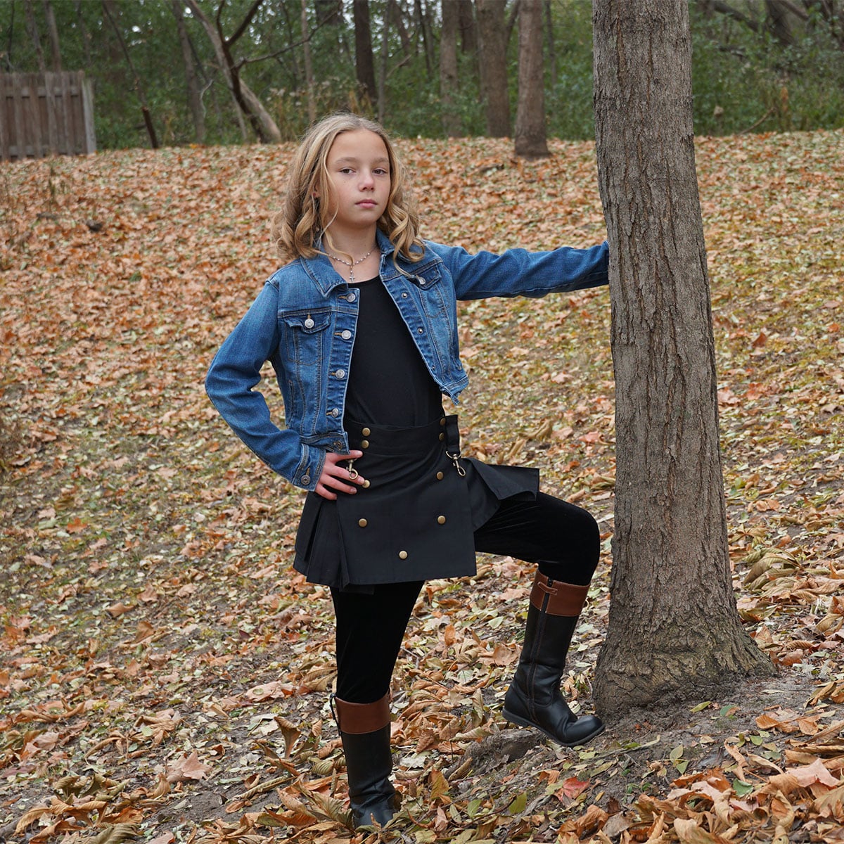 A girl is leaning against a tree in the fall, wearing Wilderness Kilts for Kids.