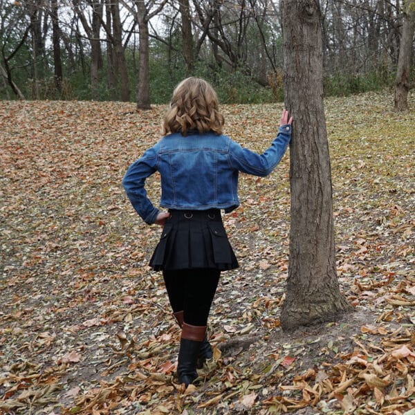 A girl wearing Wilderness Kilts for Kids standing next to a tree in a wooded area.