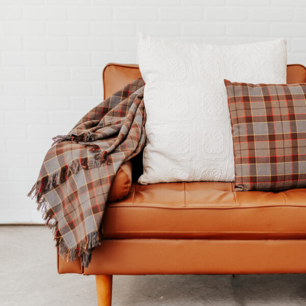 A brown leather couch with plaid pillows and an OUTLANDER lambswool tartan scarf draped gracefully on one side.