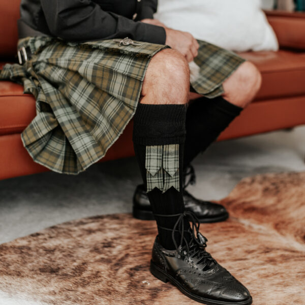 A man in a plaid kilt sitting on a couch, showcasing the Kilt and Accessories Bundle - Homespun Wool-Blend.