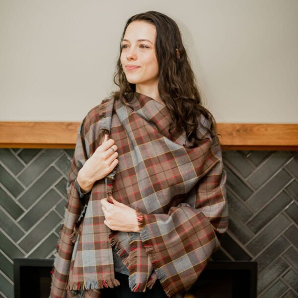 A woman wearing a plaid OUTLANDER Shawl Poly/Viscose Tartan in front of a fireplace.
