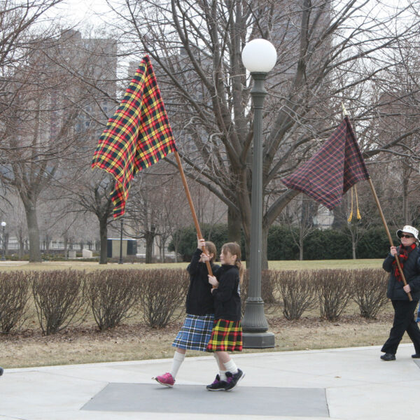 A group of people wearing Medium Ancient Kid Kilts walking down a sidewalk with flags, some holding ancient symbols aloft. The kilts add a vibrant touch to the procession.