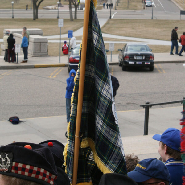 A group of Medium Ancient Kid Kilt-wearing people, including kids, standing on a sidewalk holding a flag.