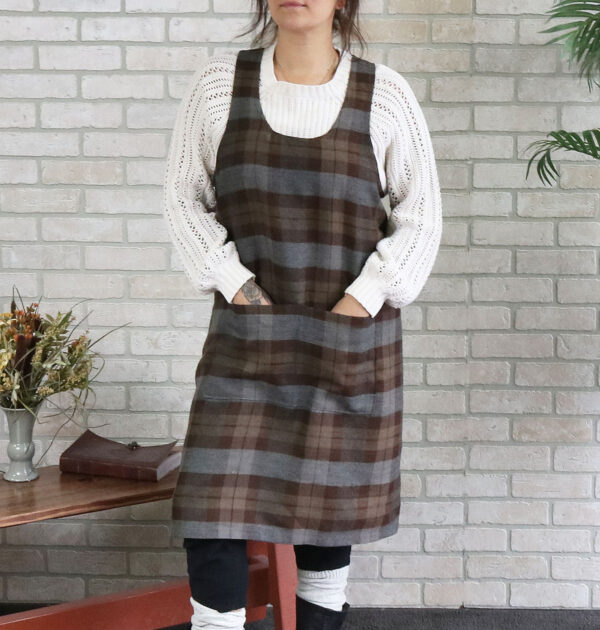 A woman wearing a Tartan Cross Back Apron - Homespun Wool Blend standing in front of a brick wall.