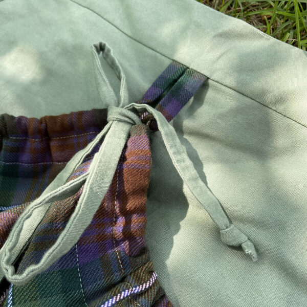 A Tartan Gathering Apron - Homespun Wool-Blend with a plaid tie on the ground.