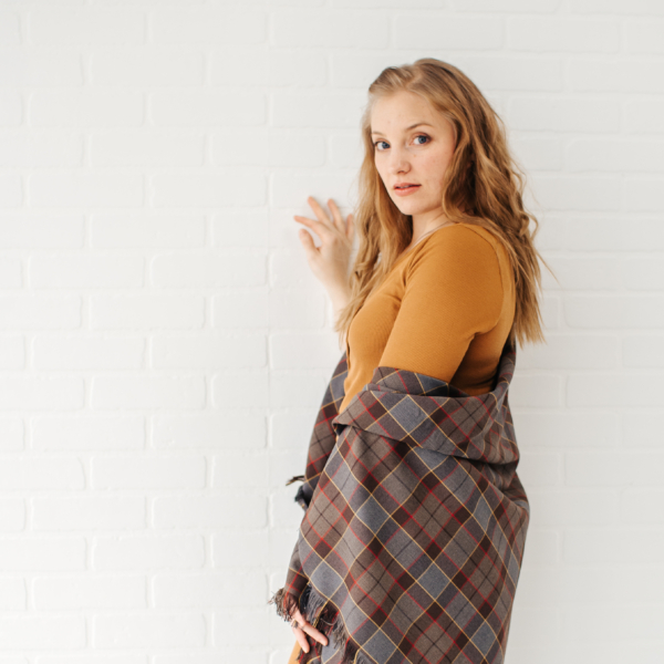 A young woman in an OUTLANDER Wrap Premium Lambswool Tartan leaning against a brick wall.